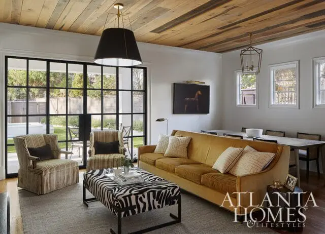 Living room with a mustard velvet sofa, zebra print ottoman, striped armchairs, and a wood plank ceiling. Large black-framed windows look out onto a patio.
