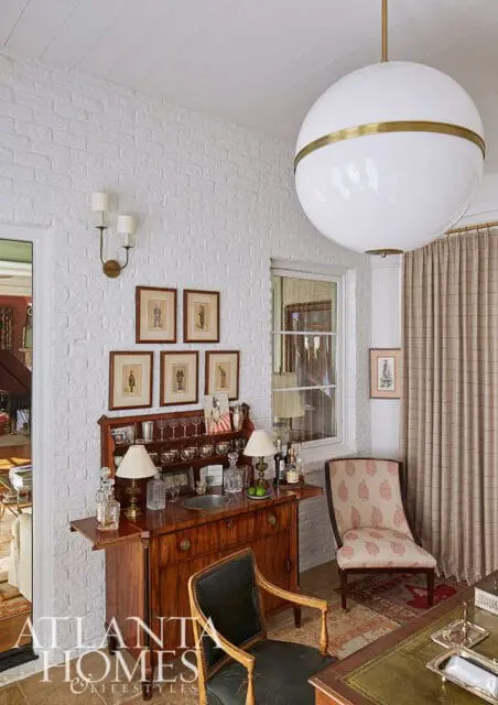 White brick walls in a room with a large globe pendant light, a wooden bar cabinet stocked with glassware and bottles, and two chairs.
