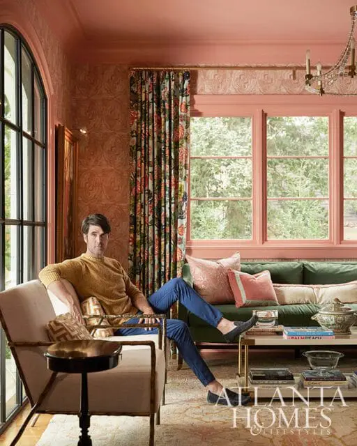 Man in yellow sweater and blue pants sitting in a chair in a room with coral walls, floral curtains, and a green sofa.