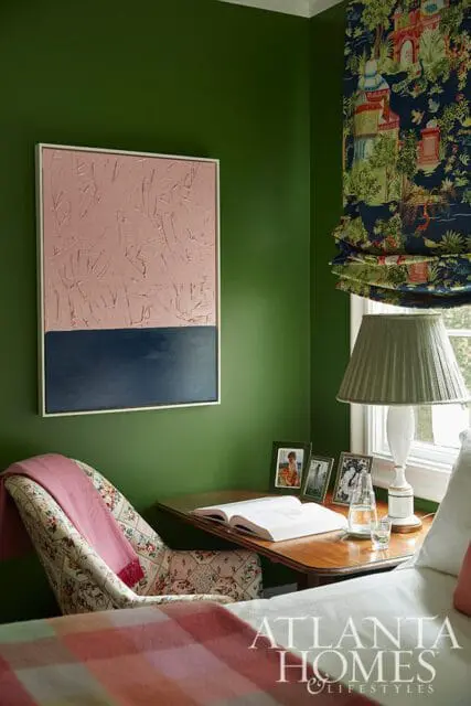 Green walls with abstract art, floral armchair with pink throw, wooden desk with open book, framed photos, and a lamp under a patterned Roman shade.
