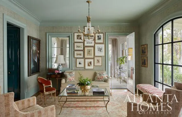 Sage green sitting room with textured wallpaper, a cream sofa, glass coffee table, gallery wall, and arched window overlooking greenery.
