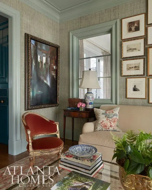Sitting room with textured sage green walls, a red velvet armchair with gold trim, a neutral sofa with floral pillows, and a collection of framed art above a window.