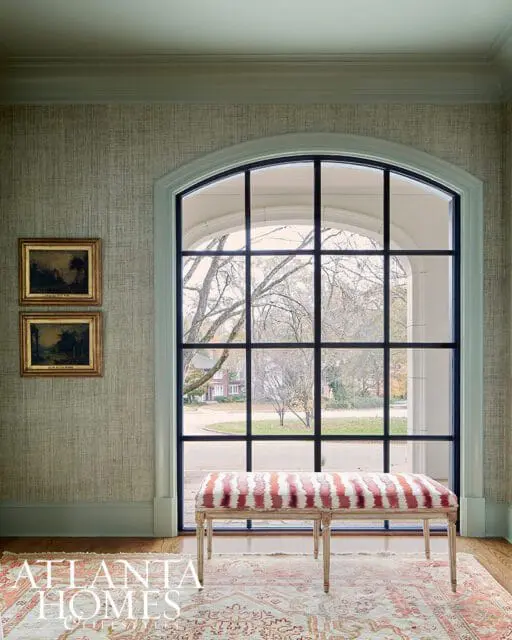Sitting room with sage green textured wallpaper, a large arched window with black mullions, a striped bench, and an oriental rug.
