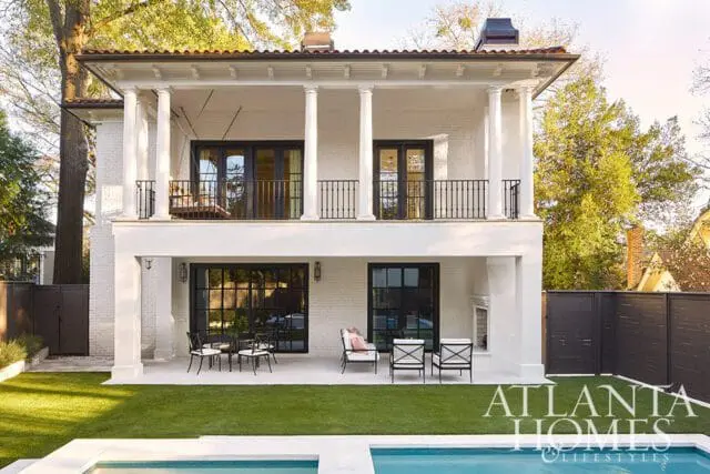 White brick house with a covered balcony supported by columns, black-paned doors and windows, and a tiled roof. A patio with outdoor furniture overlooks a pool and lawn.