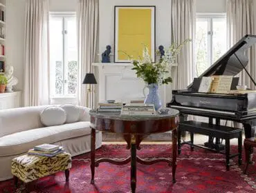 Living room with a white curved sofa, grand piano, round marble-top table, fireplace with yellow artwork, and built-in bookshelves.