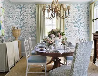 Dining room with blue and white floral wallpaper, a round wooden dining table set for a meal, and upholstered chairs with patterned seat cushions.