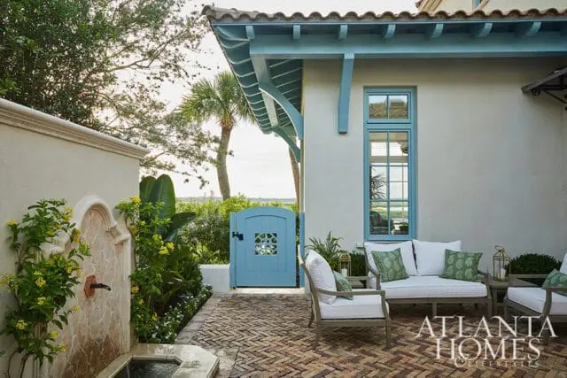 Courtyard with a stone fountain, herringbone brick patio, light-colored stucco walls, and a light blue arched gate. Outdoor seating with white cushions and green patterned pillows is visible.
