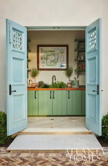 Light blue double doors open to a potting shed with green cabinetry, a wooden countertop, sink, and framed garden plan on the wall.