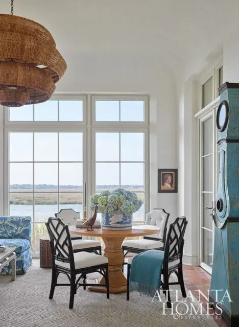 Dining area with a round wood pedestal table, four black and white chairs, a blue and white floral vase filled with hydrangeas, and a large wicker chandelier hanging overhead. Large windows offer a view of a body of water and marshland.
