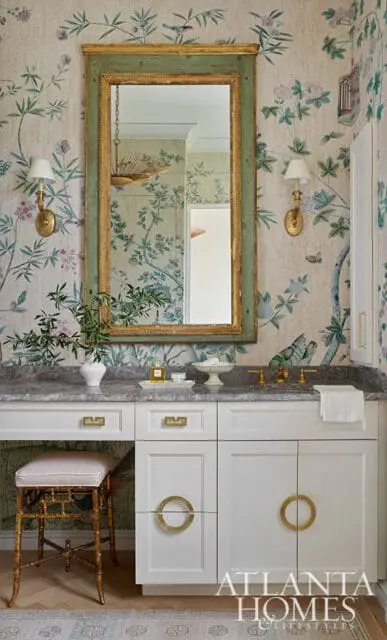 Powder room with Chinoiserie wallpaper, a green and gold framed mirror, white vanity with marble countertop, brass hardware, and a gold bamboo stool.