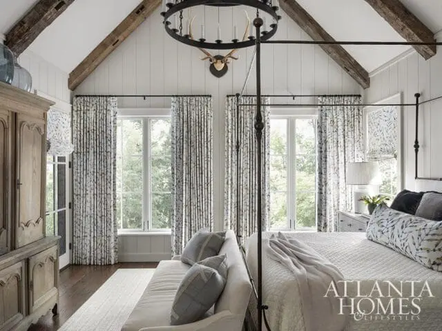 Bedroom with vaulted ceiling, exposed beams, and antler chandelier. Features a four-poster bed, patterned curtains, and a rustic armoire.