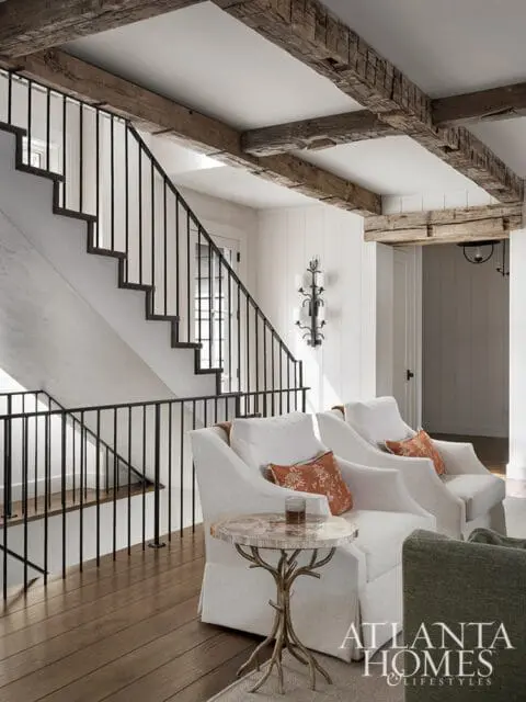 Living room with white slipcovered armchairs, rustic wooden beams on the ceiling, a black metal staircase railing, and a small side table with a petrified wood top.