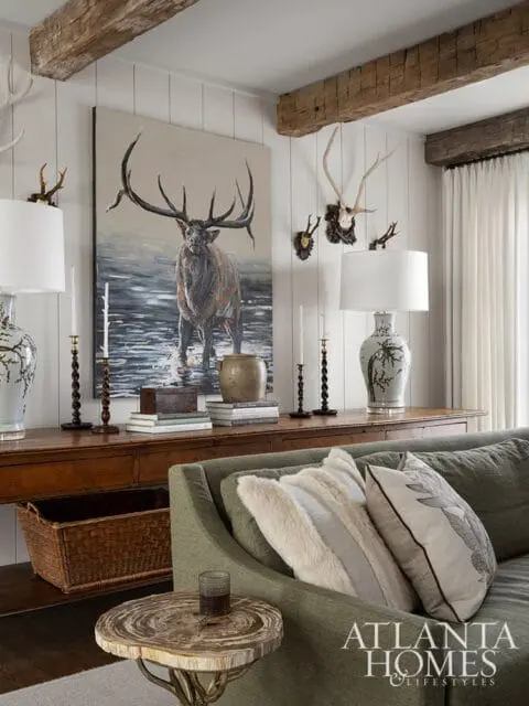Living room with rustic wooden beams, a large painting of an elk, mounted antlers, a wooden console table, and a green sofa with decorative pillows.