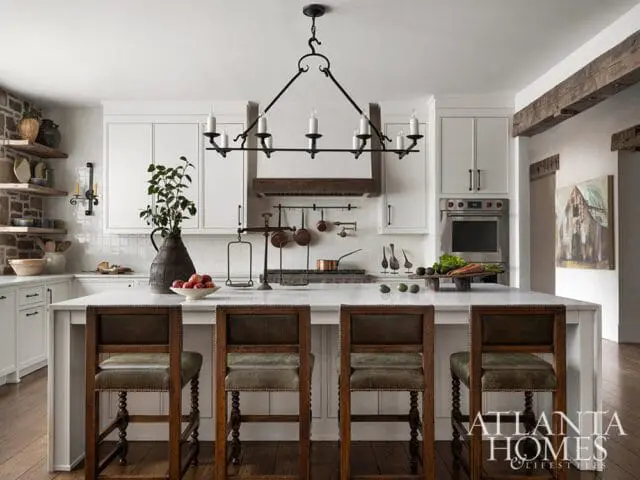 White kitchen with a marble island, four leather bar stools, stone accent wall, and a wrought iron chandelier.