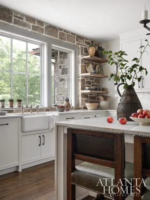 Rustic stone accent wall in a white kitchen with a farmhouse sink, marble island, and wooden bar stools.