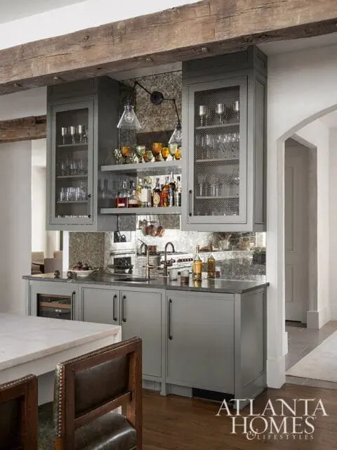 Gray bar cabinet with glass-fronted doors displaying glassware and bottles, a mirrored backsplash, and a wine fridge. Exposed wooden beams are visible overhead.