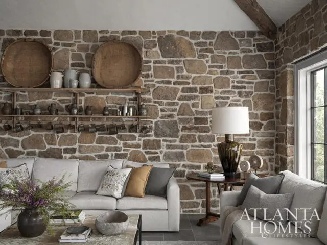 Living room with a stone accent wall, light gray sectional sofa, wooden shelves with pottery and baskets, and a round side table with a lamp.