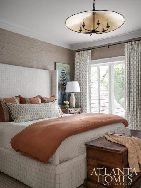 Bedroom with a white upholstered bed, textured wallpaper, patterned curtains, and a large brass chandelier. An antique wooden chest sits at the foot of the bed.