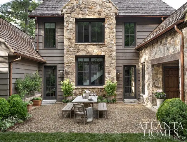 Rustic stone and wood-sided home with a gravel courtyard featuring a picnic table and benches, surrounded by manicured greenery.