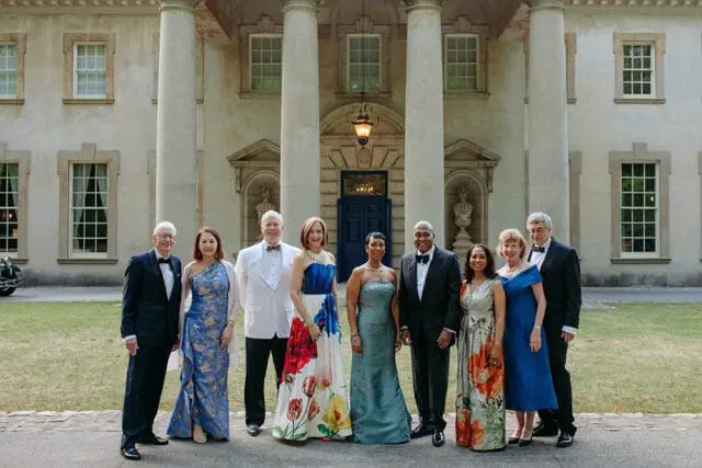 Group of eight people in formal attire standing in front of a neoclassical building with large columns and a blue door.