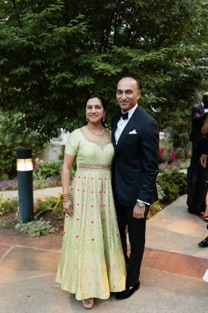 A couple poses for a photo outdoors. The woman wears a light green dress with pink diamond patterns and gold jewelry. The man wears a black tuxedo with a bow tie.