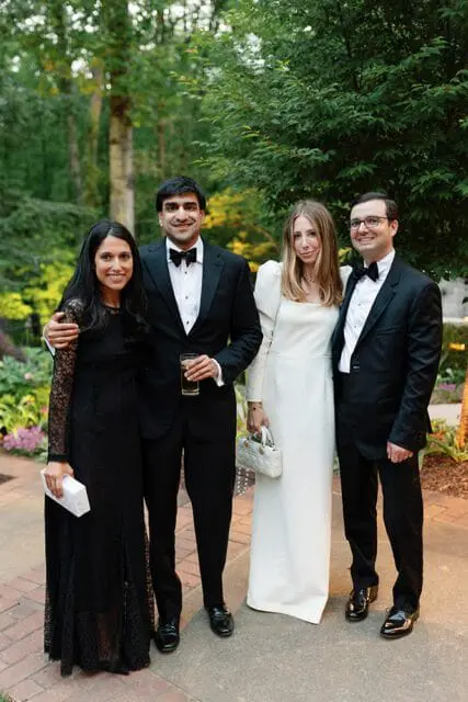 Two couples dressed in formal attire stand outdoors with trees and greenery in the background.