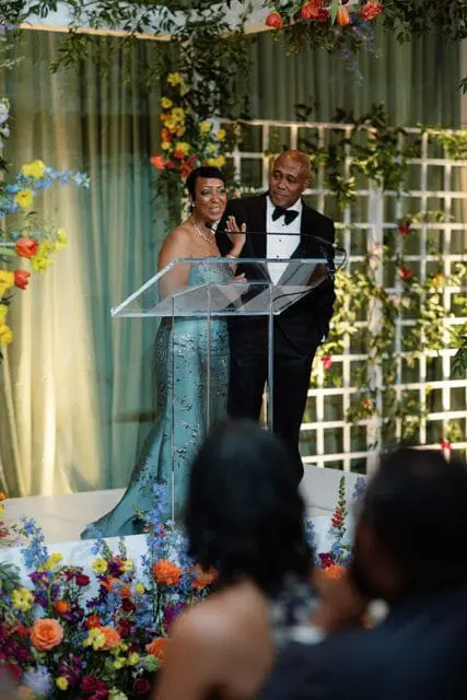 A woman in a strapless blue sequined gown and a man in a tuxedo stand at a podium, speaking at an event.