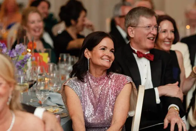 Woman in a sparkly pink and silver dress smiles while seated at a formal dinner table next to a man in a tuxedo with a red bow tie.
