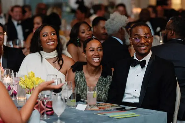 Three people smiling at a formal event. A woman in a white dress is on the left, a woman in a green sparkly dress is in the middle, and a man in a tuxedo is on the right.