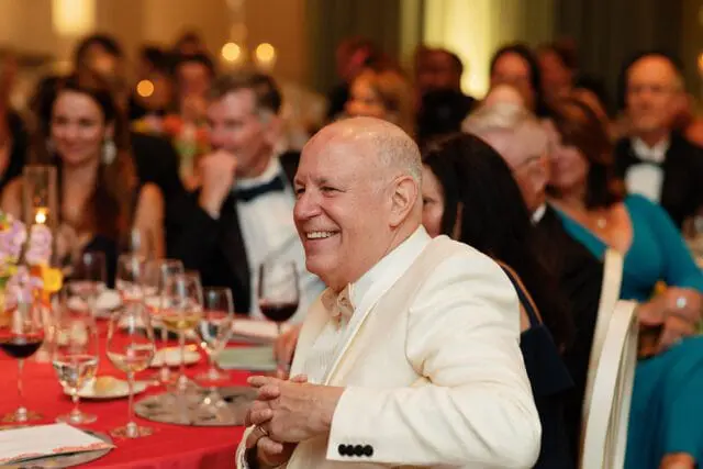 Man in a cream-colored suit smiling at a formal dinner event with other guests blurred in the background.