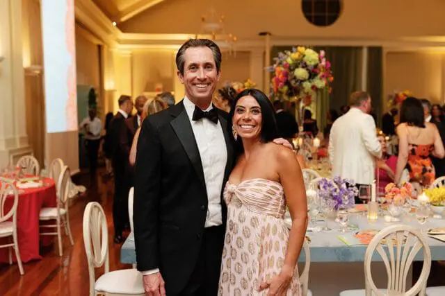 Man in a black tuxedo and woman in a pink strapless dress with gold pattern smile at a formal event with tables set for dinner in the background.
