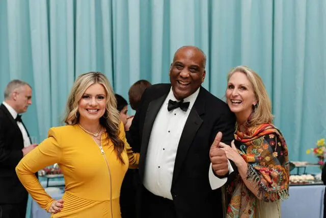 Three people smiling at a formal event. A woman in a yellow dress is on the left, a man in a tuxedo is in the middle giving a thumbs up, and a woman in a colorful scarf is on the right.
