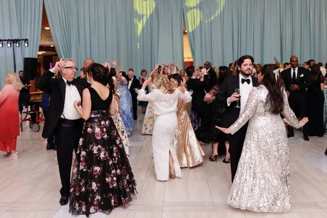 People in formal attire dancing at an event with light blue curtains in the background and yellow lighting projected on them.