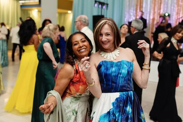 Two women smiling at a formal event. The woman on the left wears a floral dress and the woman on the right wears a blue strapless gown and a statement necklace.