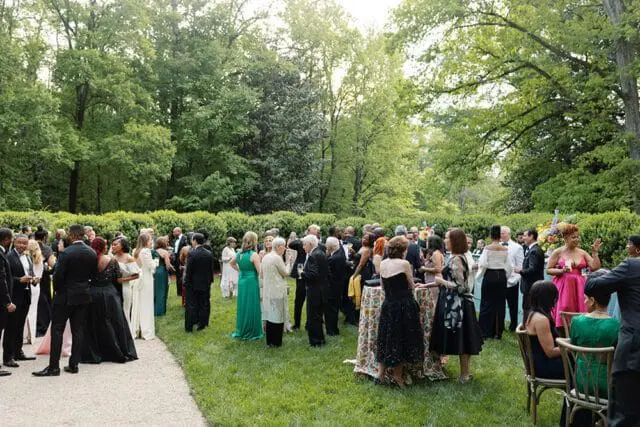 A large group of people in formal attire mingle on a grassy lawn surrounded by lush trees and manicured hedges during an outdoor event.