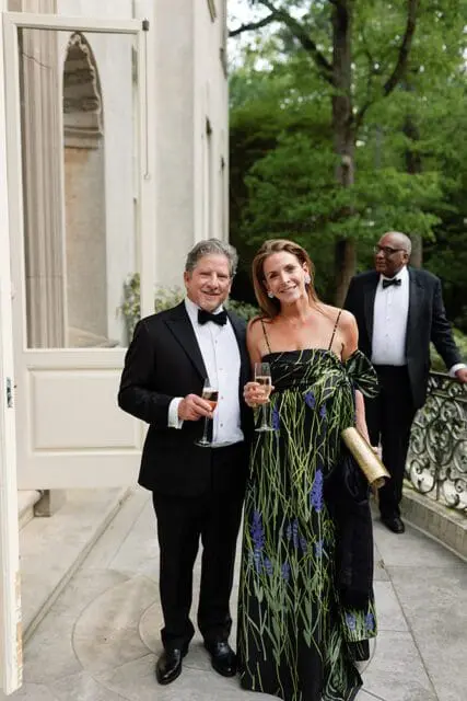 Man in a tuxedo holding a champagne flute and woman in a floral dress holding a champagne flute, with another man in a tuxedo in the background.