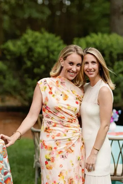 Two women smiling at the camera outdoors. The woman on the left wears a cream-colored dress with a floral print in shades of pink, orange, and yellow. The woman on the right wears a cream-colored sleeveless dress with a ribbed texture.