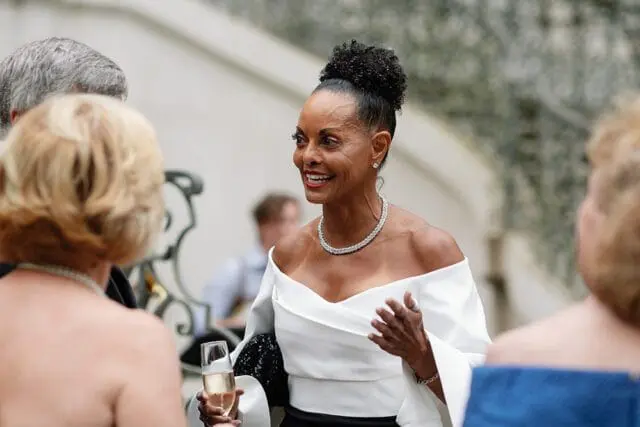 Woman with dark curly hair in a bun, wearing a white off-the-shoulder top and diamond necklace, holding a champagne flute and talking to others at an outdoor event.
