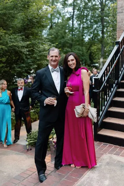 Couple in formal attire posing outdoors with a brick staircase and lush greenery in the background. The man wears a black tuxedo with a bowtie, and the woman wears a vibrant pink gown.