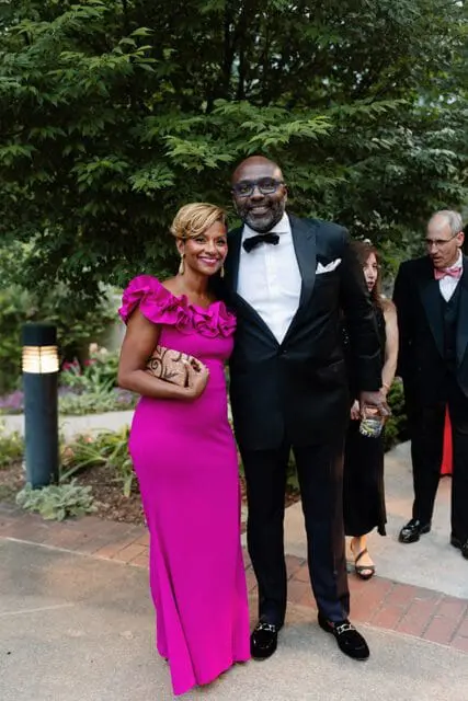 A Black couple smiles at the camera, the woman in a bright pink off-the-shoulder gown with ruffled trim and the man in a black tuxedo with a bow tie and glasses.
