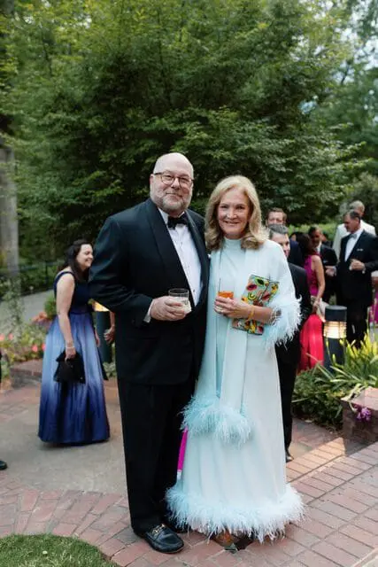 Man in tuxedo and woman in light blue feathered dress holding drinks at an outdoor event