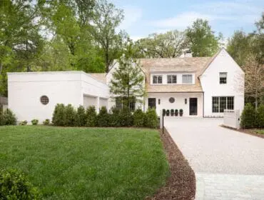 White brick house with a light wood shingle roof, black-framed windows, and a circular window on the garage. A gravel driveway leads to the entrance, flanked by manicured hedges and a lush green lawn.