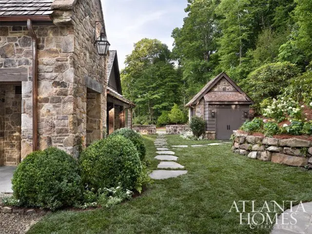 Stone home with wood shake roof and copper gutters, leading to a stone pathway. A small stone garage with a wood shake roof sits on a hill surrounded by lush greenery.
