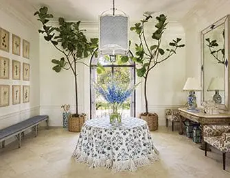 Foyer with a round table covered in a blue and white floral tablecloth, large potted plants, a long bench, and a mirror above a console table.