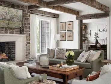 Living room with stone fireplace, rustic wood beams, green sofas, and botanical prints on the wall. A view into a white kitchen is visible in the background.