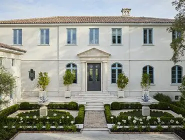 Mediterranean-style stucco house with a tile roof, light blue-framed windows, and a formal garden with manicured hedges and white flowers.