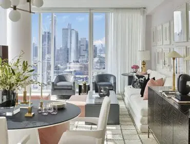 Living room with large windows overlooking a city skyline, featuring a white sofa, grey armchairs, a dark wood sideboard, and a round dining table with a floral arrangement.
