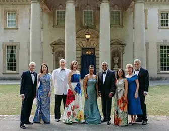 Group of ten people in formal attire standing on a paved walkway in front of a large building with white columns and a dark blue door.