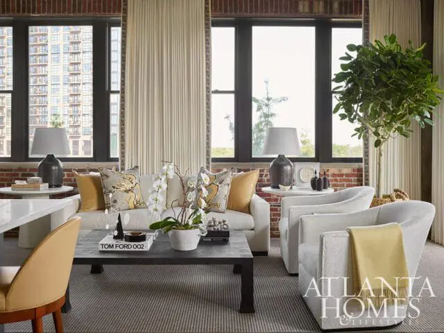 Living room with exposed brick walls, cream sofa with patterned pillows, two white armchairs with a yellow throw, and a dark coffee table with books and a plant.