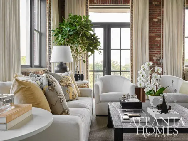 Living room with white sofa, white armchairs, dark wood coffee table, and exposed brick walls. A large plant and white orchids add greenery.